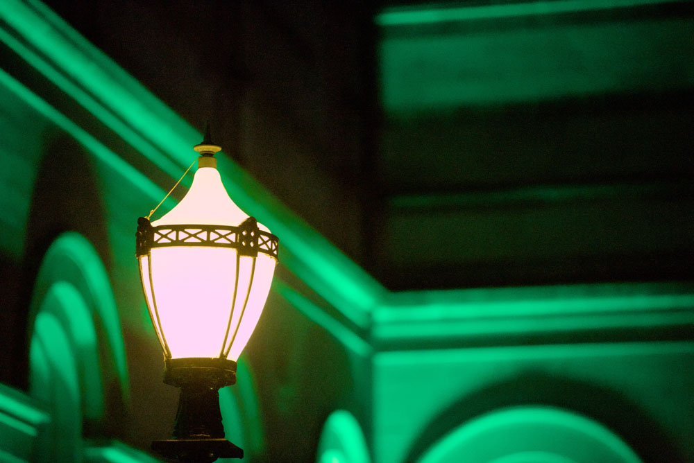 A photograph of an old-fashioned streetlamp taken at 200mm focal length. The head of the streetlamp is in focus, with the facade of a green-lit building blurred in the background.
