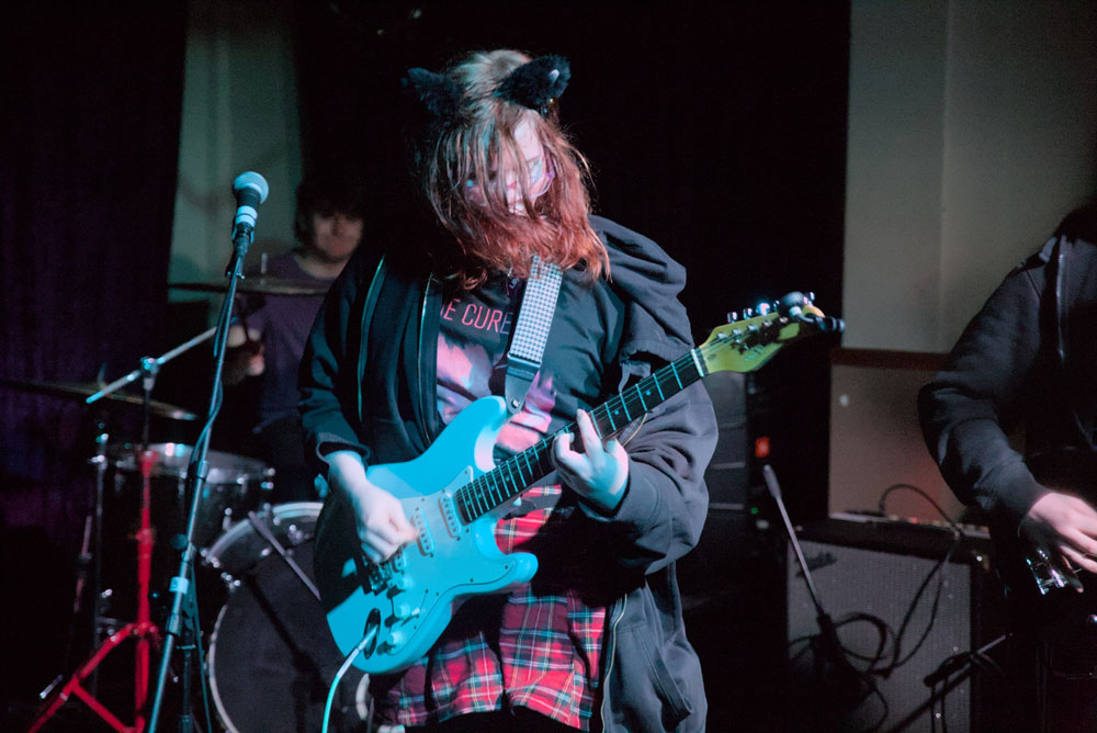 A photo of a girl playing an electric guitar. She wears a black hoodie, a The Cure shirt, a red skirt and cat ears.