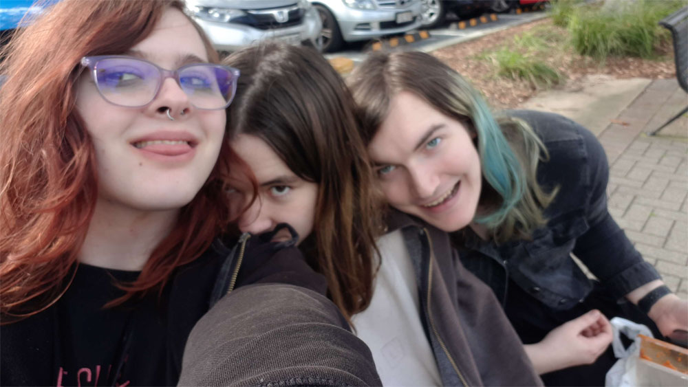 Three girls sitting on a park bench. On the left is the guitarist from previous images, in the middle is the photographer from the 24/200mm comparison, and on the right is a girl with dyed blue hair.